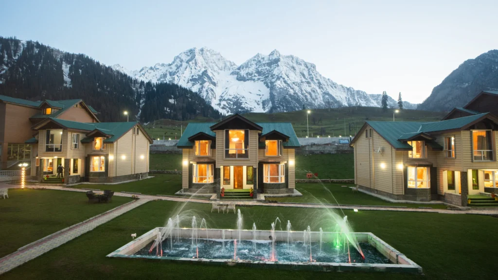 Sindh resort cottages in Sonamarg with illuminated rooms and a garden fountain, set against snow-covered Himalayan mountains at dusk.